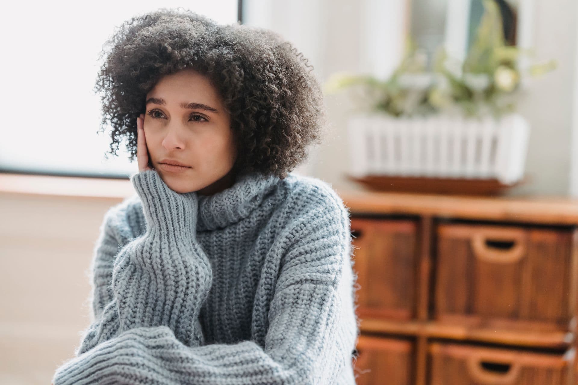 A woman in a blue sweater resting her cheek on her hand, gazing away with a worried expression
