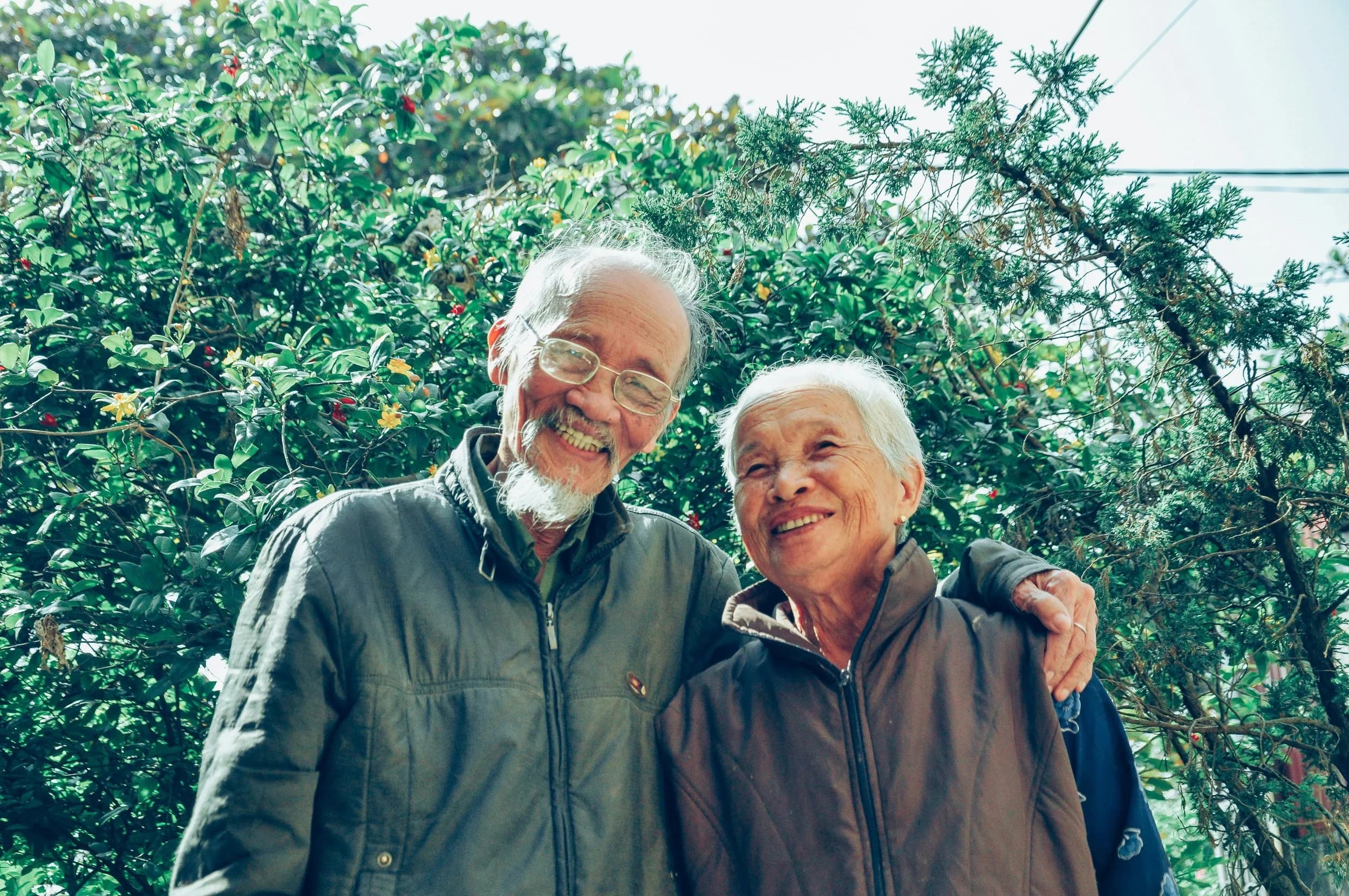 An older couple smiling together outdoors, his arm around her shoulder