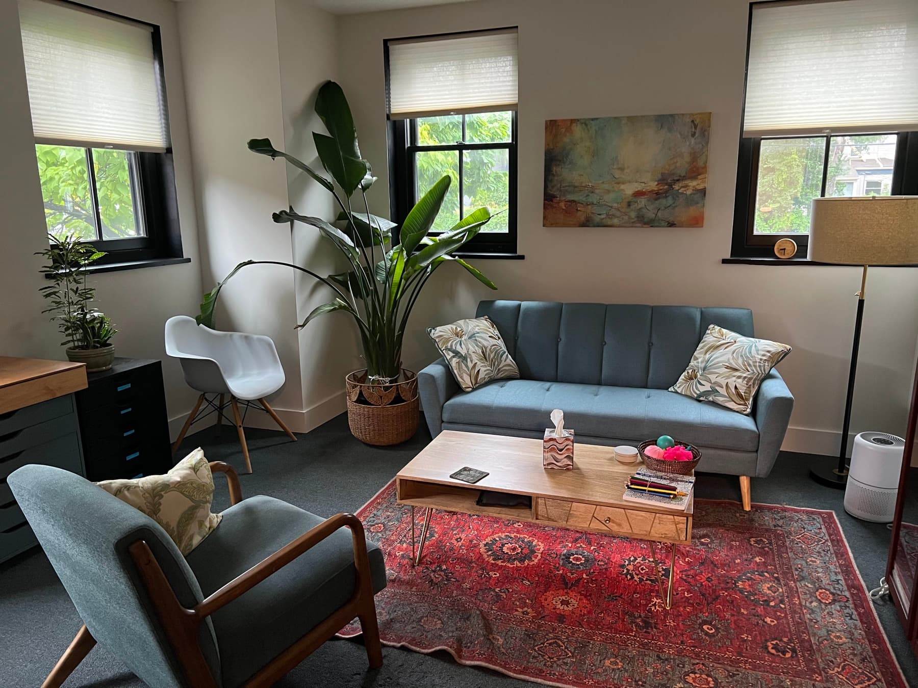A bright therapy room with a blue sofa, an armchair, plants, and large windows
